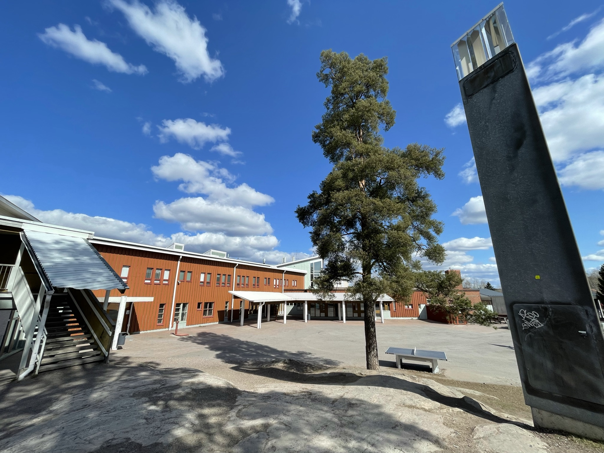 Kuva Nöykkiön koulun pihalta. Etualalla näkyy pihan metallitorni, taustalla alfalttikentän takana punatiilinen koulurakennus.